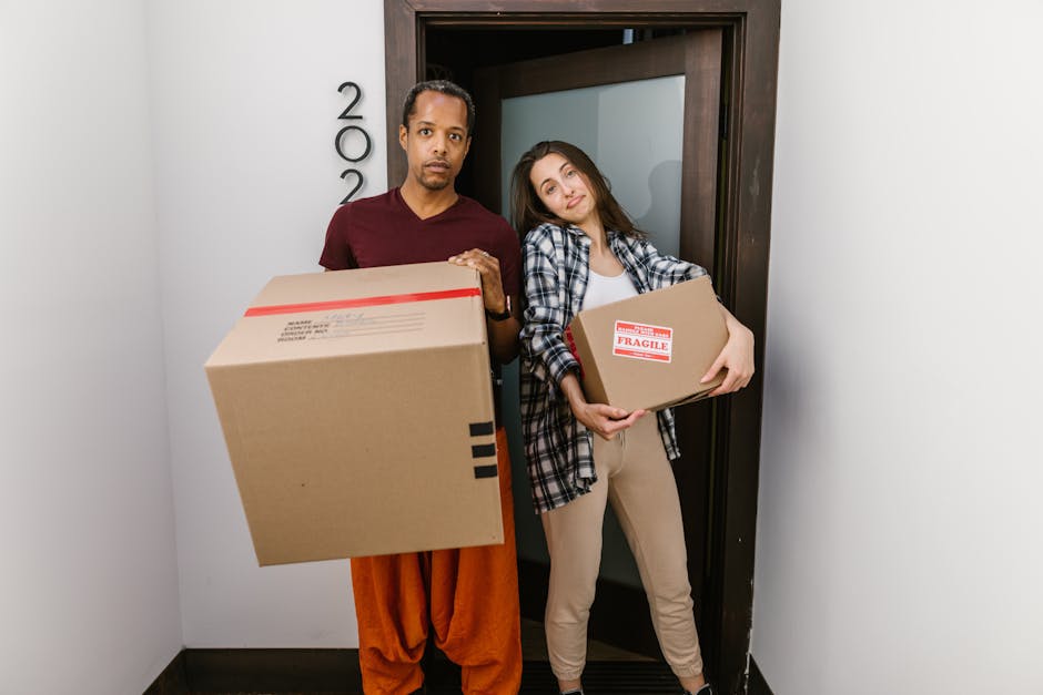 A man and a woman stand inside a doorway of a residential building, both holding cardboard boxes used for home relocation. The man, wearing a maroon t-shirt and orange trousers, holds a large box in front of him, while the woman, dressed in a plaid shirt and beige pants, holds a smaller box labeled 'Fragile' with red tape and handling instructions. They are positioned on a grey tiled floor next to a white wall with black house numbers and a dark wooden door frame. The lighting is bright and natural, indicating an indoor environment during packing and moving preparations. This scene reflects the process of furniture transport and packing during a house removal, typical of professional removals services such as those provided by Man With a Van Maida Hill, ensuring a smooth home relocation.