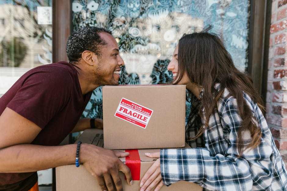 Two movers from Man With a Van Maida Hill are engaged in a home relocation task outside a building with large glass windows and a brick wall. The man on the left, with dark curly hair and wearing a ma