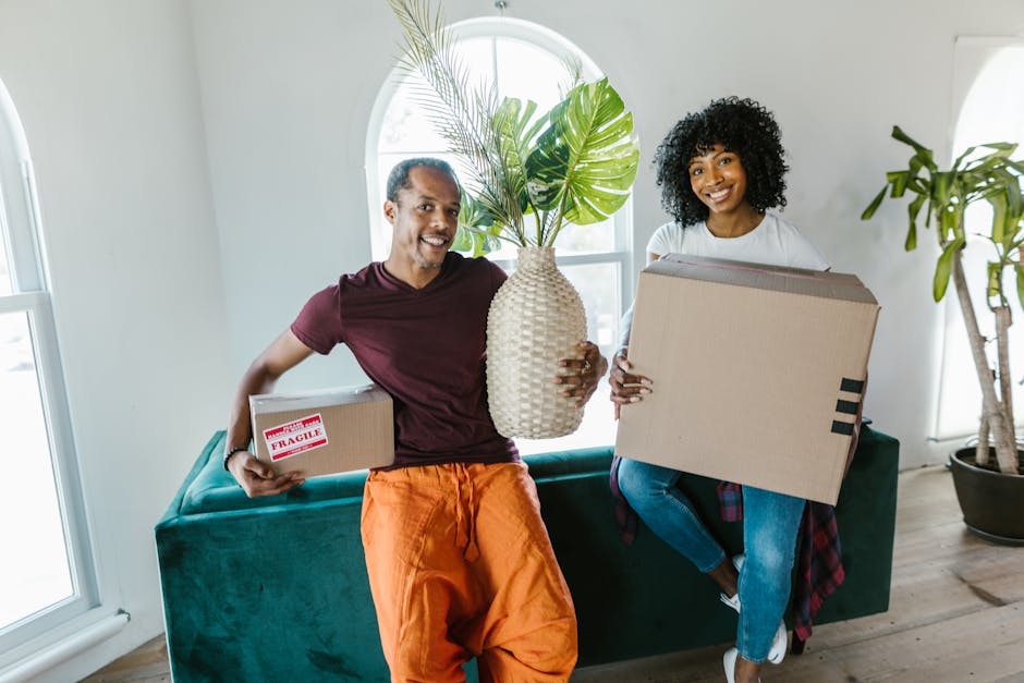 Two movers from Man With a Van Maida Hill are engaged in a home relocation task outside a building with large glass windows and a brick wall. The man on the left, with dark curly hair and wearing a ma