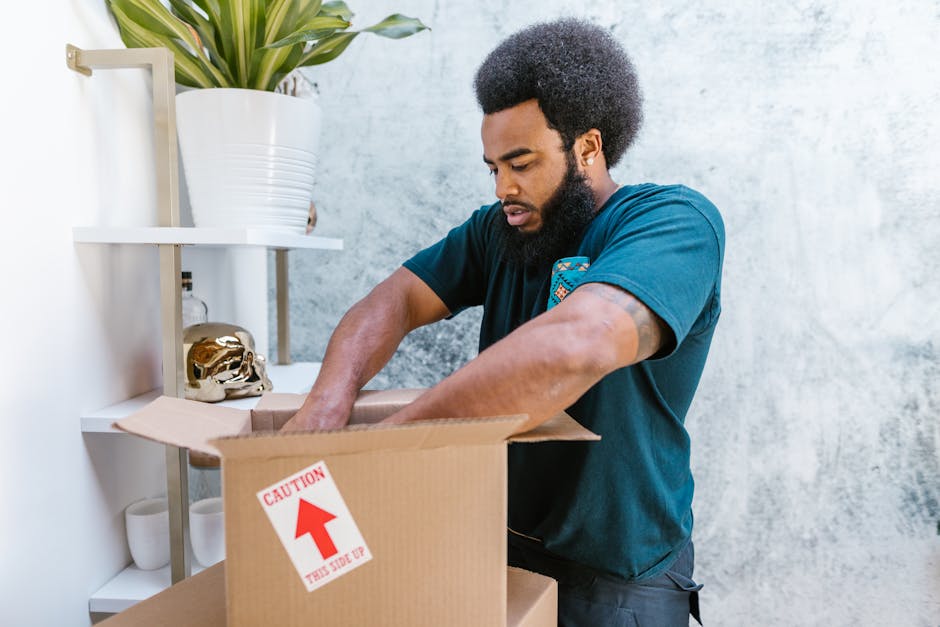 A man with an Afro hairstyle and beard, wearing a teal t-shirt, is seen packing a cardboard moving box in an indoor space. He is standing next to a white shelving unit that holds a large white pot with a green leafy plant, a gold-colored decorative object, and three white cups. The man is placing items inside the open box, which has a red and white caution label on the side indicating 'This Side Up.' The background features a light-colored, textured wall, and the scene is lit with natural lighting, depicting a home relocation or packing process as part of furniture transport and moving preparations, consistent with services offered by Man With a Van Maida Hill.