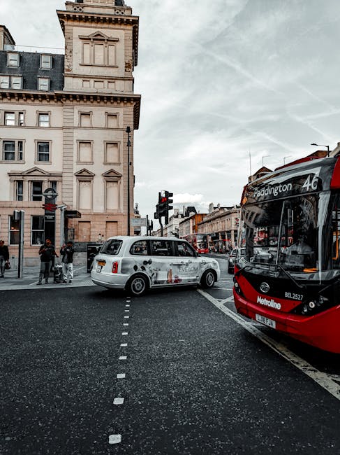 The image shows a busy street scene near Paddington Station in Maida Hill, with a red and black public bus stopped on the right side of the road. A white, vintage-style vehicle is positioned perpendicular to the curb, appearing to be involved in a home relocation or furniture transport process managed by Man With a Van Maida Hill. The vehicle's rear doors are open, and several cardboard boxes, wrapped furniture, and packing materials are visible inside or nearby, indicating packing and moving activities. The boxes are made of brown cardboard, some with plastic wrapping or padding for protection. A few people are standing on the sidewalk, observing or assisting with the loading process. The setting includes classic Victorian-style buildings with detailed facades and large windows, under a cloudy sky with some contrails. The scene captures both the logistics of moving in an urban environment and the ongoing street traffic, serving as context for house removals and transport services near Paddington Station.