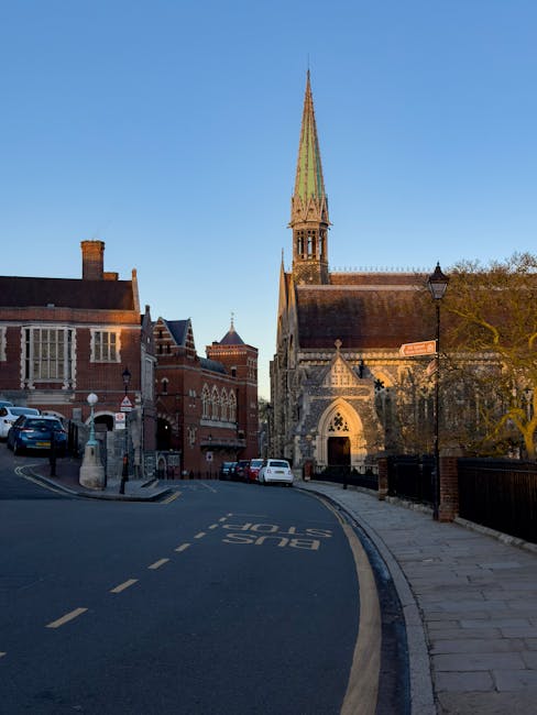 A narrow street in Maida Hill with a gentle curve, featuring parked cars along the curb and a bus stop marking painted on the asphalt. Streetlights and trees line the sidewalk, which is paved with stone slabs. In the background, a historic church with a tall, pointed spire and detailed stonework dominates the scene, while surrounding buildings with brick facades and traditional architecture are visible. The sky is clear and blue, suggesting daytime with natural light illuminating the area. This setting represents a typical residential and community environment where Man With a Van Maida Hill might assist with house removals, furniture transport, and home relocation services, particularly considering access and parking conditions for loading and unloading. Material details such as the vehicles, street signage, and the church’s stone features are accurately depicted, supporting visual understanding of a typical moving operations environment.