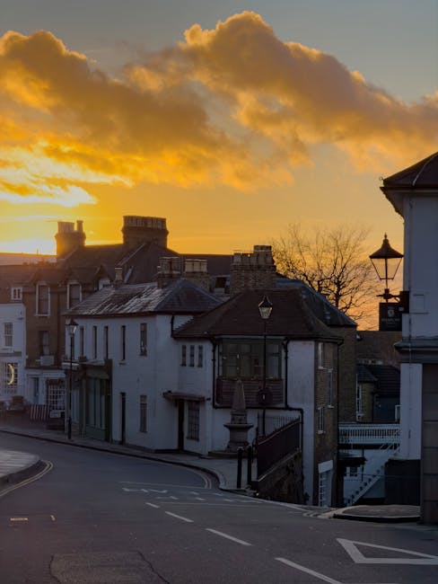 Photograph of a residential street at sunset in Maida Hill, showcasing a row of house facades constructed from brick and plaster with pitched roofs, chimneys, and multiple windows. The street is quiet with a gentle curve, and features lampposts and a black metal railing along the pavement. In the foreground, a collection of cardboard boxes, wrapped furniture, and moving blankets are arranged on the pavement, suggesting preparations for home relocation. Visible within the scene are a white van parked nearby, partially obscured by the buildings, indicating a typical moving vehicle used by Man With a Van Maida Hill. The warm glow of the setting sun accentuates the buildings and the sky is filled with orange and yellow hues with scattered clouds. This scene illustrates the logistics and environment associated with furniture transport and packing during a house removal process in the Maida Hill area.