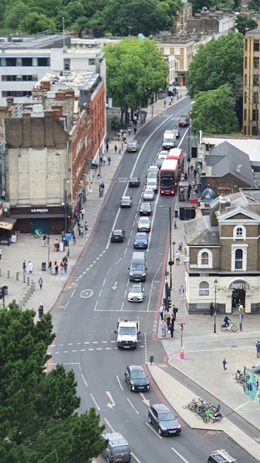 A narrow street in Maida Hill with a gentle curve, featuring parked cars along the curb and a bus stop marking painted on the asphalt. Streetlights and trees line the sidewalk, which is paved with stone slabs. In the background, a historic church with a tall, pointed spire and detailed stonework dominates the scene, while surrounding buildings with brick facades and traditional architecture are visible. The sky is clear and blue, suggesting daytime with natural light illuminating the area. This setting represents a typical residential and community environment where Man With a Van Maida Hill might assist with house removals, furniture transport, and home relocation services, particularly considering access and parking conditions for loading and unloading. Material details such as the vehicles, street signage, and the church’s stone features are accurately depicted, supporting visual understanding of a typical moving operations environment.