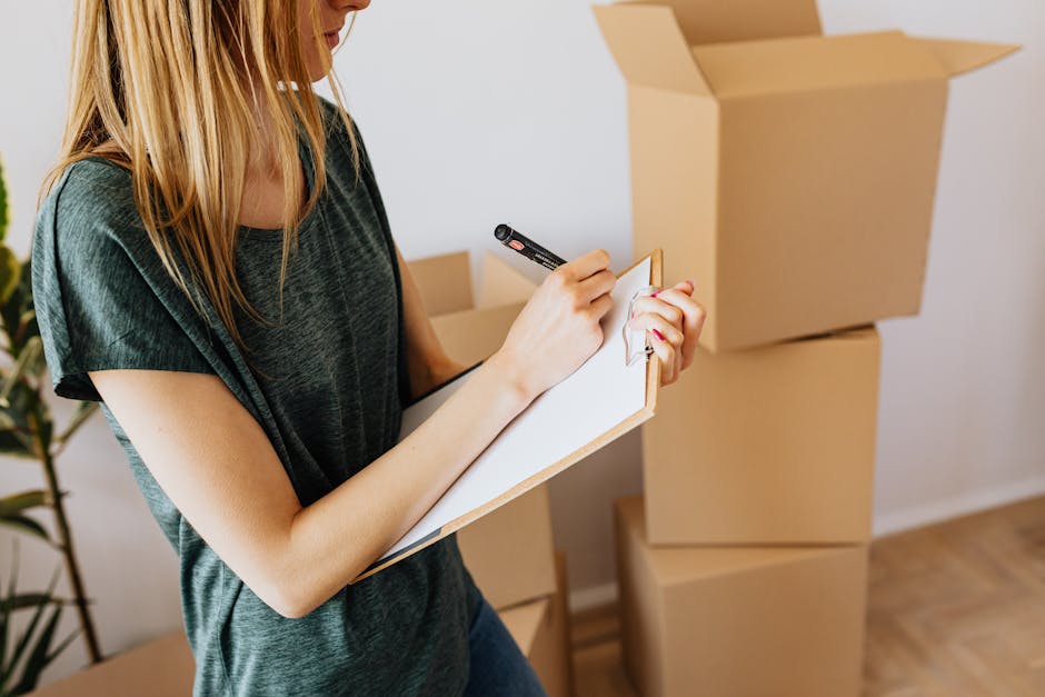 A woman with long blonde hair, wearing a dark grey T-shirt, is indoors surrounded by several large, open cardboard moving boxes stacked on the floor. She is holding a clipboard in her left hand and writing with a black marker in her right hand. The boxes appear to contain packing materials and household items, indicating a home relocation process. In the background, there is a white wall and part of a potted plant. The scene captures a moment of organizing or checklist-taking related to house removals, with natural lighting highlighting the packing and moving preparation, consistent with the services provided by Man With a Van Maida Hill as seen on the Kilburn Lane flat removals checklist webpage.