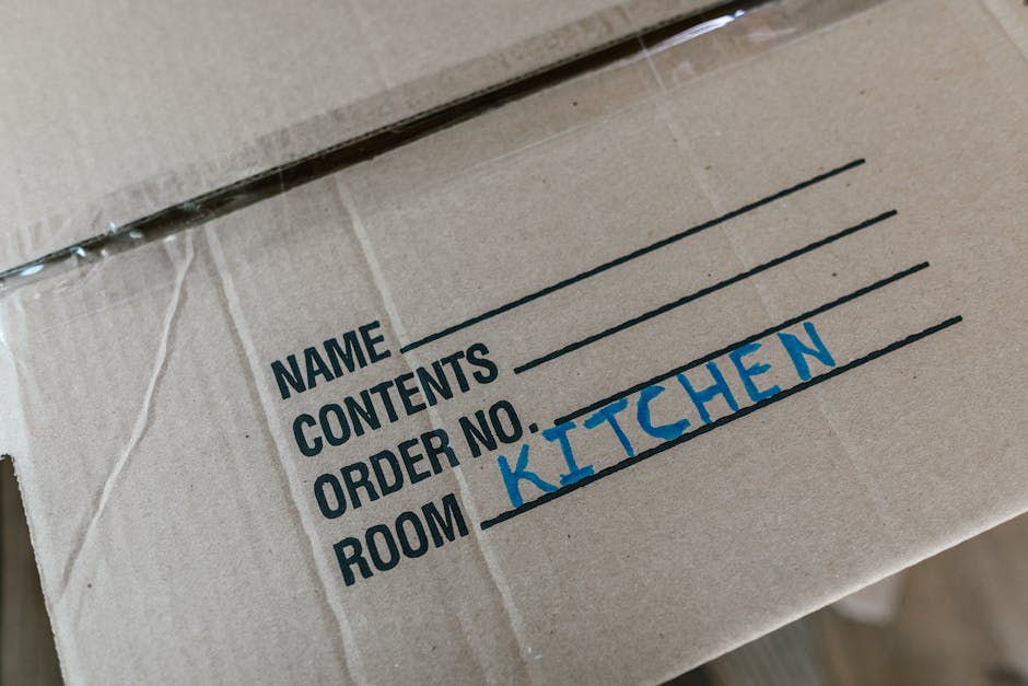 Close-up view of a cardboard moving box with a handwritten label indicating the contents. The label shows the room as 'KITCHEN' written in blue marker, with sections for the sender's name, contents, and order number visible above. The box appears to be inside a property, likely in a hallway or near a doorway, ready for loading onto a van. The box is sealed with clear packing tape, and the background includes other packed boxes, indicating an ongoing home relocation process. The image emphasizes proper packing and labelling as part of furniture transport and packing and moving services, with regional context related to Maida Hill house removals as provided by Man With a Van Maida Hill.