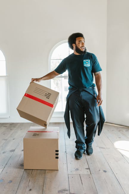 A woman with long blonde hair, wearing a dark grey T-shirt, is indoors surrounded by several large, open cardboard moving boxes stacked on the floor. She is holding a clipboard in her left hand and writing with a black marker in her right hand. The boxes appear to contain packing materials and household items, indicating a home relocation process. In the background, there is a white wall and part of a potted plant. The scene captures a moment of organizing or checklist-taking related to house removals, with natural lighting highlighting the packing and moving preparation, consistent with the services provided by Man With a Van Maida Hill as seen on the Kilburn Lane flat removals checklist webpage.
