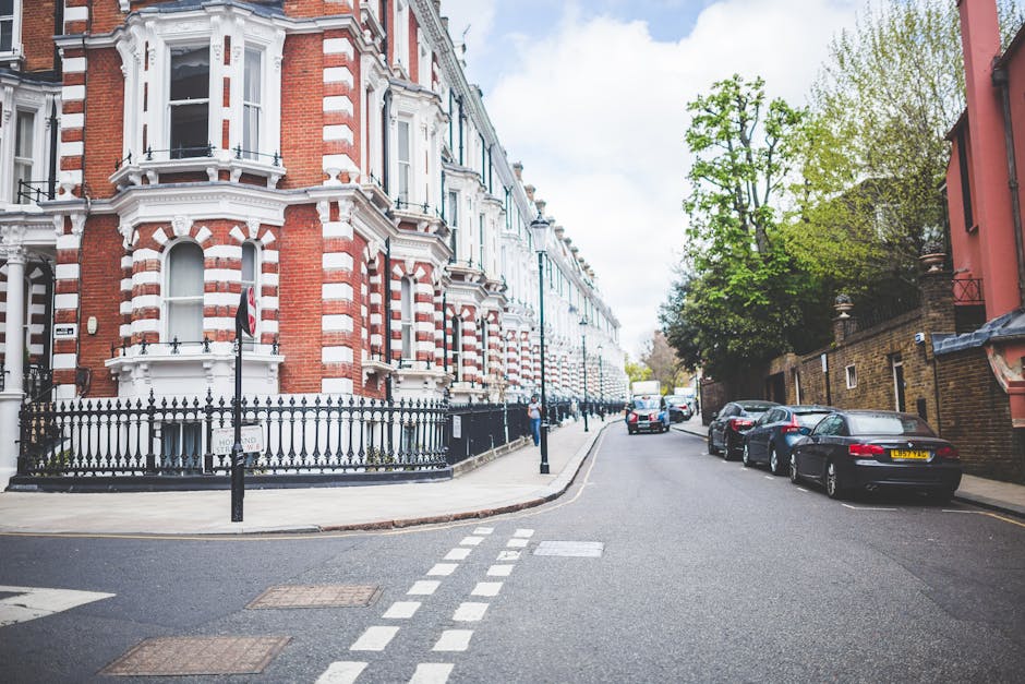 A wide view of a residential street in Maida Hill during daytime, featuring Victorian-style terraced houses with red and white brick facades, decorative window moldings, and black wrought iron railings along the pavement. Several cars are parked along the right side of the street, including a black sedan and a silver hatchback, with additional vehicles visible further down the road. The pavement area adjacent to the houses is partly blocked by black bollards, and the sky is partly cloudy with patches of blue. To the right, there is a row of trees with green foliage, providing some natural shading. This scene captures the typical urban environment where house removals and furniture transport activities take place, with loading zones and street access considerations relevant to Maida Hill council removal rules and street permit guidance, as handled by Man With a Van Maida Hill.