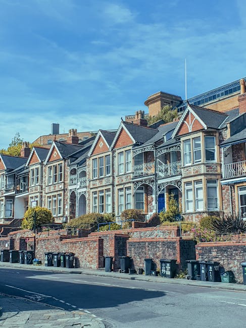 Image depicts a row of Victorian-style terraced houses with ornate balconies and large bay windows, situated on a residential street under a bright blue sky. The houses are built with brick and feature decorative white trim around the windows and balconies. In the foreground, a low brick wall with steps and small landscaped gardens separates the houses from the pavement. Several black wheelie bins are lined up along the pavement, indicating waste collection. The street appears calm without visible vehicles or pedestrians, suggesting a quiet neighbourhood. This scene is representative of urban residential areas where house removals or furniture transport might occur, with the houses potentially requiring street permits or adherence to local removal rules, as outlined in Maida Hill council regulations. Occasionally, man with a van services like Man With a Van Maida Hill may assist in home relocation or moving logistics in such areas, involving careful loading and transport of household items.