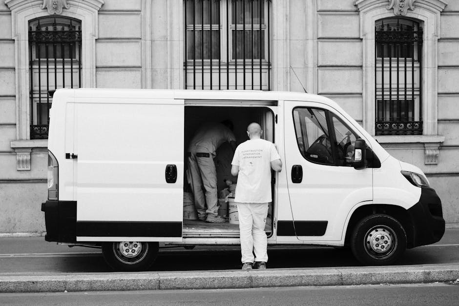 A white panel van with its side sliding door open is parked on a city street directly in front of a stone building with barred windows. Inside the van, several movers are engaged in a home relocation task, with one person bent over stacking or adjusting items while another is partially visible further inside. Outside the van, a man wearing a white T-shirt and light trousers stands with his back to the camera, observing or assisting the loading process. The van is positioned on the pavement next to a curb, and the background features a historic-style building with classic architectural features. The scene is illuminated by natural daylight, underscoring the process of furniture transport and packing and moving activities typical of professional removals carried out by Man With a Van Maida Hill, supporting efficient house removals and logistics.