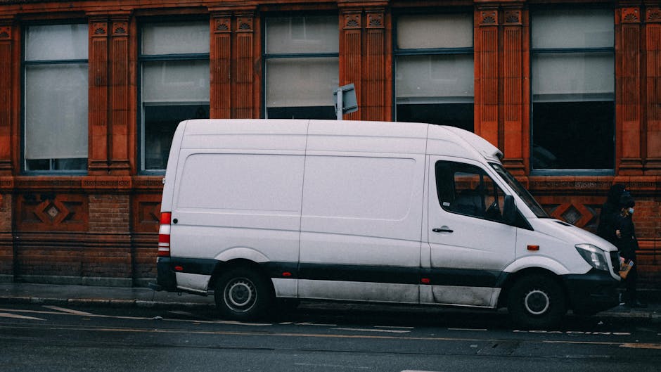 A white delivery van with a streamlined body and black rubber trim along the lower sides is parked parallel to the curb on a city street. The van's windows include a large windshield, a side window for the driver, and a smaller window in the side door, with the vehicle positioned close to the brick building behind it. The background features a red brick building with decorative architectural details around the windows, which are partially covered with blinds or curtains. The van is situated on a paved road with double yellow lines indicating parking restrictions, and a woman is walking past the van on the sidewalk, which is separated from the street by a curb. The scene captures a typical urban environment suitable for home relocation or furniture transport services, with the van ready for loading or unloading items such as boxes, furniture, or packaging materials involved in house removals or moving logistics, as coordinated by Man With a Van Maida Hill.