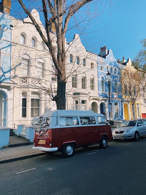 A white panel van with its side sliding door open is parked on a city street directly in front of a stone building with barred windows. Inside the van, several movers are engaged in a home relocation task, with one person bent over stacking or adjusting items while another is partially visible further inside. Outside the van, a man wearing a white T-shirt and light trousers stands with his back to the camera, observing or assisting the loading process. The van is positioned on the pavement next to a curb, and the background features a historic-style building with classic architectural features. The scene is illuminated by natural daylight, underscoring the process of furniture transport and packing and moving activities typical of professional removals carried out by Man With a Van Maida Hill, supporting efficient house removals and logistics.