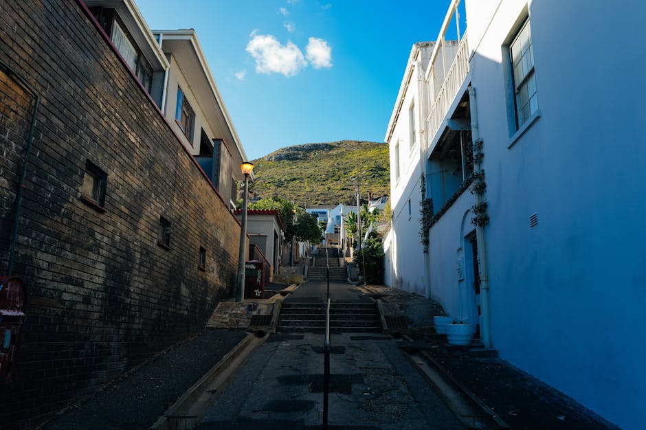 A steep residential street in Maida Hill with narrow stairs leading up between white and brick buildings, some with balconies and greenery, under a clear blue sky. A streetlamp is situated along the sidewalk, which is adjacent to the steps. The photograph captures the outdoor environment during daytime, highlighting the challenging access for furniture transport and home relocation activities. The buildings are multi-story with a variety of window styles, and vegetation is visible on some structures. This setting illustrates common issues faced during removals for properties with limited stairwell space, where careful planning of furniture handling and logistics is essential, as often managed by [COMPANY_NAME], Man With a Van Maida Hill, for efficient packing and moving procedures.