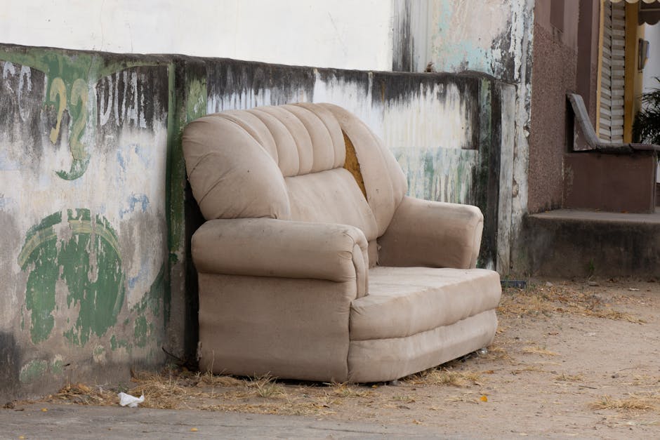 An old beige fabric armchair with padded armrests and a slightly worn appearance, positioned outdoors against a weathered wall with peeling paint and graffiti in shades of green and blue. The chair is placed on a rough, dirt ground with scattered leaves and a small piece of trash nearby. Behind the chair, a concrete step and a brown building entrance are visible, indicating an exterior setting close to a residential property in Maida Hill. The scene captures part of a home relocation or furniture transport context, with the chair awaiting disposal or removal, which is relevant to safe furniture disposal rules for Westminster removals as addressed by Man With a Van Maida Hill.