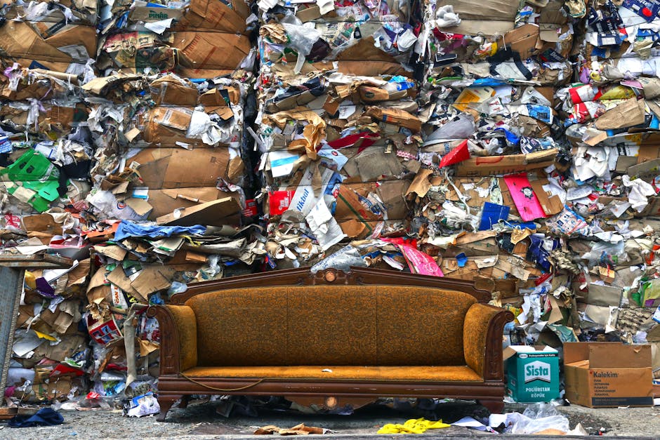 A vintage wooden sofa with patterned upholstery is positioned in front of a large wall of tightly packed recyclable paper, cardboard, and plastic waste. The sofa is situated outdoors on a concrete surface, near a curb, with some packing materials and boxes visible nearby, including a cardboard box labeled 'Sista' on the right. The scene suggests waste collection or disposal, with a focus on the conditions involved in furniture disposal during a home relocation or move. The background waste is organized in large bales, and there is natural daylight illuminating the scene, highlighting the textures and colors of the materials. This image illustrates the importance of proper furniture disposal and the logistical considerations in moving services offered by companies like Man With a Van Maida Hill, especially when managing bulky items during house removals in Westminster.