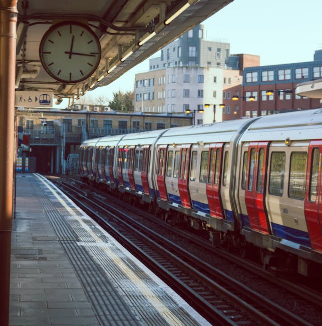 View of a train at a London underground station platform during daytime, with a large round clock hanging from the station ceiling showing the time as approximately 3:10. The train features a red, blue, and silver exterior, with multiple windows along its side. The platform is paved with rectangular tiles and has tactile paving strips near the edge for safety. In the background, there are several multi-storey buildings with modern architecture, some with large glass windows, and the sky is clear and bright. The station environment is well-lit with overhead lighting strips. This scene captures the process of home relocation or furniture transport as part of moving or packing and moving services, possibly handled by Man With a Van Maida Hill, near the busy urban area of Maida Hill, consistent with same day removals options when delays hit.