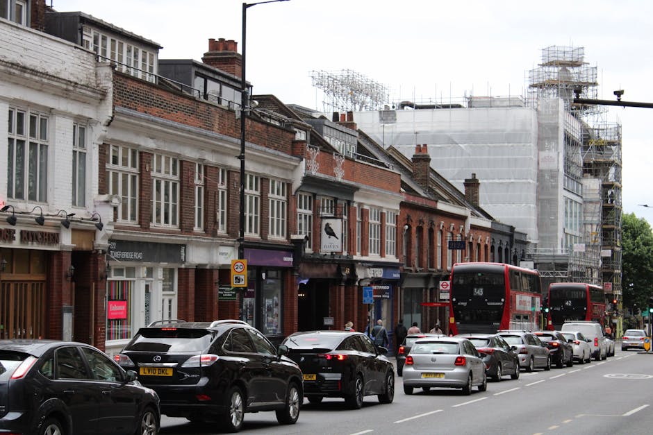 A street scene in Maida Hill showing a row of traditional brick terraced houses with large, multi-pane windows, some with white framing. The pavement in front is busy with parked cars of various models, including a black Mercedes SUV, a silver hatchback, and other compact vehicles, all lining the curb. Two red double-decker buses are stopped ahead, with route numbers 343 and 442 visible on the front, indicating local transit options, which are part of the area's public transportation network. To the right, a building under renovation is visible, covered in scaffolding and white protective sheeting, suggesting ongoing construction work. Pedestrians are seen walking along the sidewalk, some passing close to the vehicles and entrances of shops or residences. The traffic scene, combined with the urban environment, reflects typical street-level activity, including potential moving or loading processes associated with local removals services. This image naturally relates to home relocation logistics managed by firms like Man With a Van Maida Hill, highlighting the essentials of furniture transport and moving operations in a busy residential street.