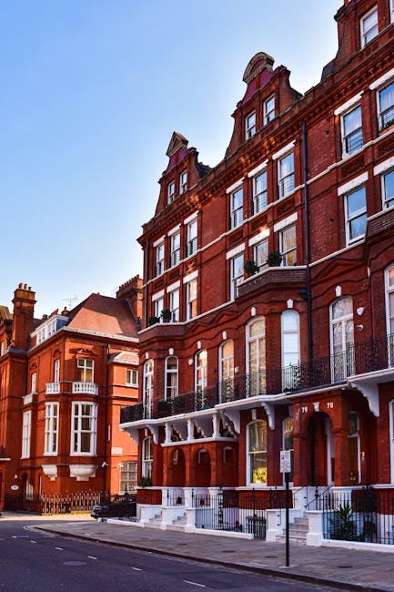 Photograph of a traditional red-brick apartment building on Maida Hill with multiple stories, large sash windows, and decorative iron railings on the balconies. The building is situated on a quiet street with a clear blue sky overhead. In the foreground, a white moving van with an open side door is parked on the street, loaded with various packing materials including cardboard boxes, plastic-wrapped furniture, and protective blankets. Several boxes and wrapped items are being carried or prepared for loading onto the van by movers not visible in the image. The scene captures the house removal process, illustrating furniture transport and packing activities associated with a home relocation, as facilitated by Man With a Van Maida Hill, serving clients in the W9 postcode area.