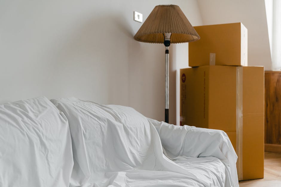 Inside a room with neutral-colored walls, three packed cardboard boxes of varying sizes are stacked near a wooden sideboard. A tall, floor-standing lamp with a beige pleated fabric shade is positioned behind the boxes, illuminating the area. In the foreground, a white sofa covered with a protective sheet is visible, indicating recent or ongoing packing and home relocation activity. The scene suggests an interior space prepared for or engaged in the process of packing items for a house removal, consistent with furniture transport and moving logistics services offered by Man With a Van Maida Hill. The lighting is natural, coming from the right side of the image, highlighting the arrangement of boxes and packing materials in preparation for a removal or moving service.