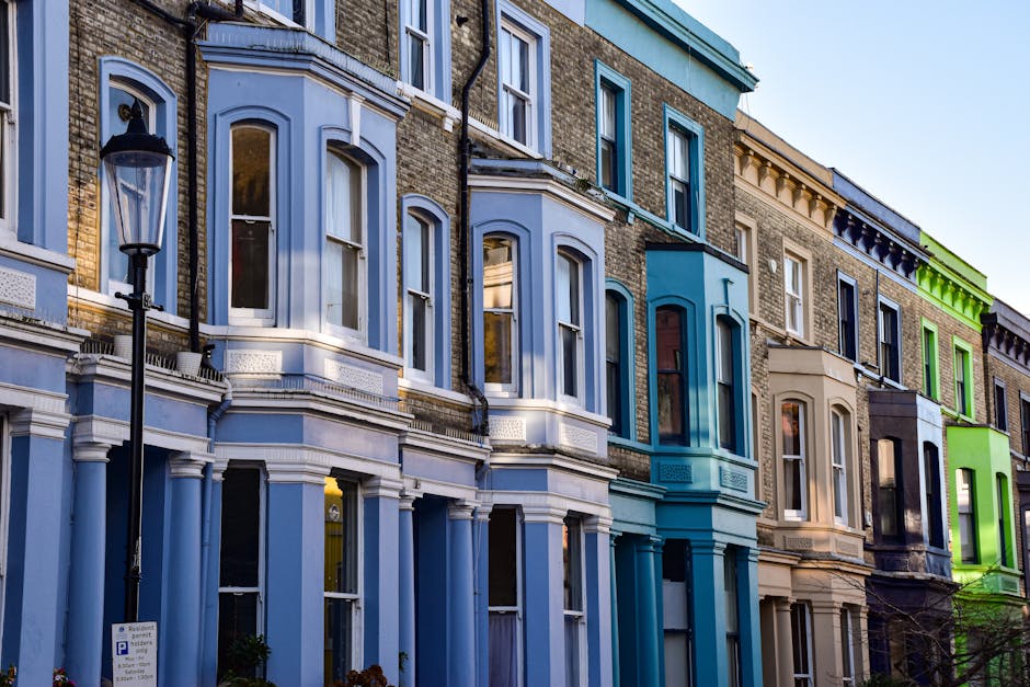 Photograph of a traditional red-brick apartment building on Maida Hill with multiple stories, large sash windows, and decorative iron railings on the balconies. The building is situated on a quiet street with a clear blue sky overhead. In the foreground, a white moving van with an open side door is parked on the street, loaded with various packing materials including cardboard boxes, plastic-wrapped furniture, and protective blankets. Several boxes and wrapped items are being carried or prepared for loading onto the van by movers not visible in the image. The scene captures the house removal process, illustrating furniture transport and packing activities associated with a home relocation, as facilitated by Man With a Van Maida Hill, serving clients in the W9 postcode area.